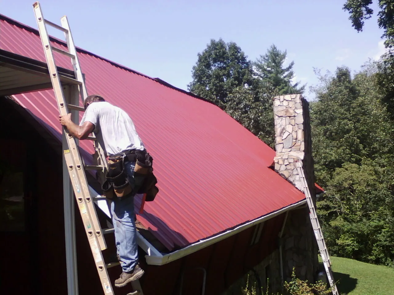 Roofer on a ladder beside a red metal roof and stone chimney — Soffit & Fascia Repair by Roofing Brothers in Foxhome